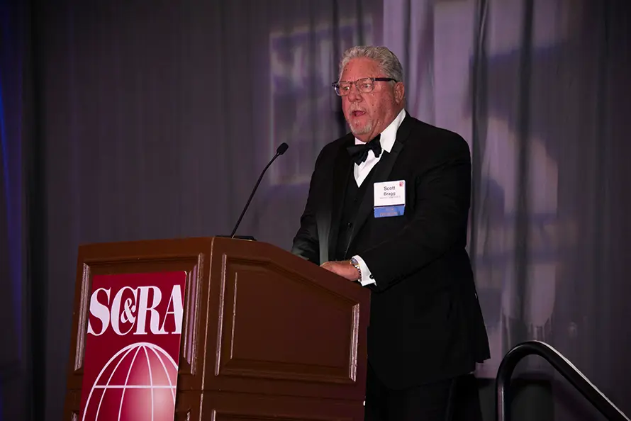 Scott Bragg speaking at a podium with the SCRA logo visible.