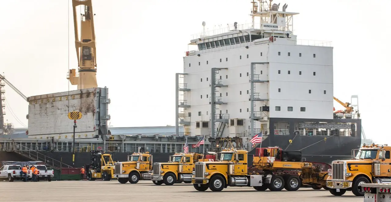 Line of heavy-duty trucks positioned near a large ship in a port or shipyard.