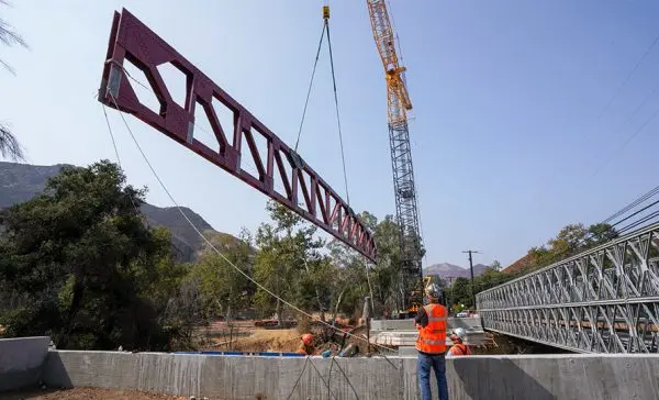 Construction workers using a crane to lift a steel beam for a pedestrian bridge.